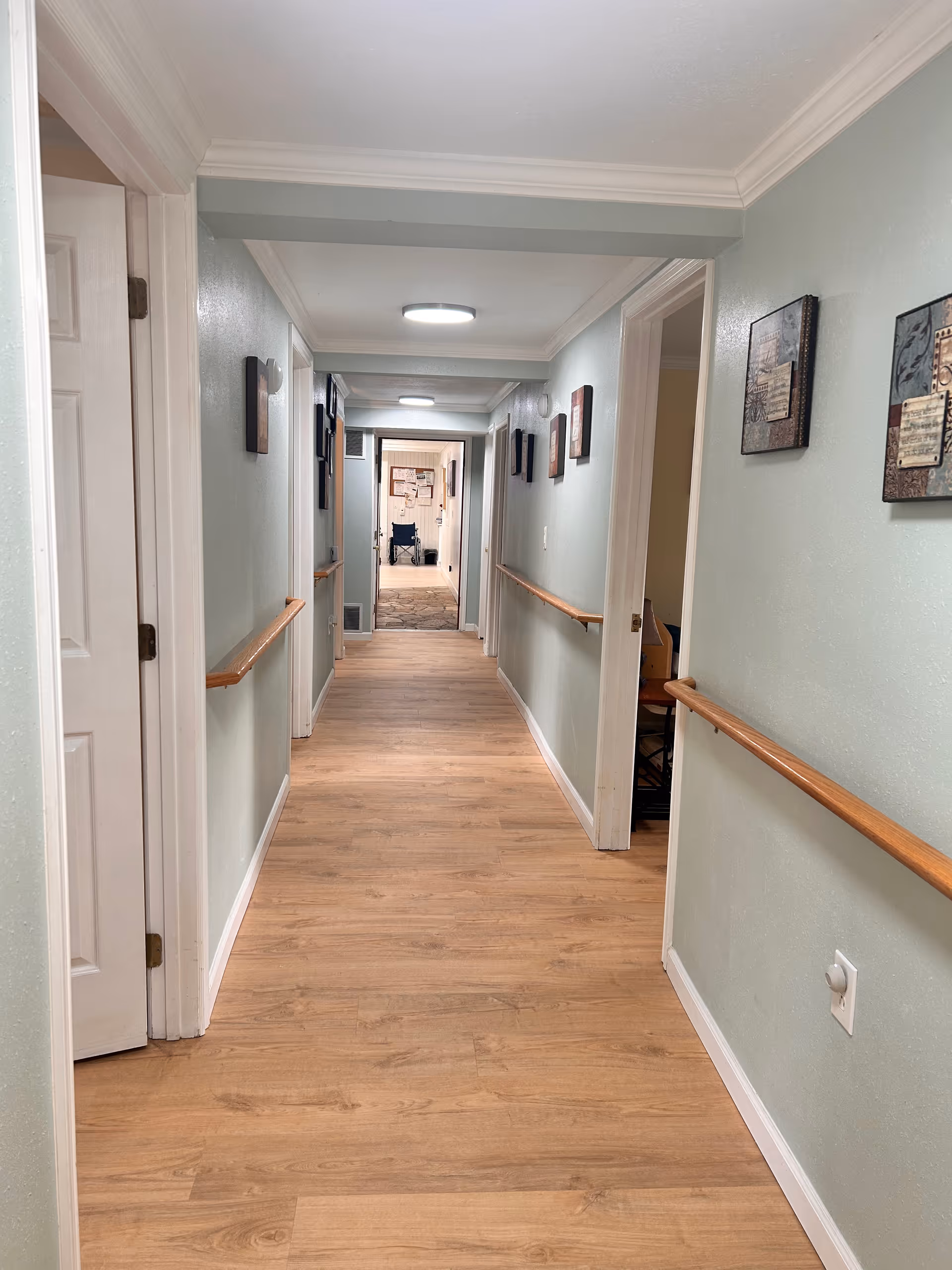 Long indoor hallway with wood-look flooring, handrails on both sides, wall art, and a wheelchair visible at the far end.