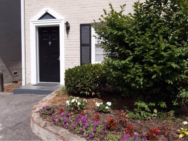 Exterior view of a building entrance with a black door, white brick walls, a black shuttered window, a wall-mounted lantern light, and a garden bed with various colorful flowers and green bushes in front.