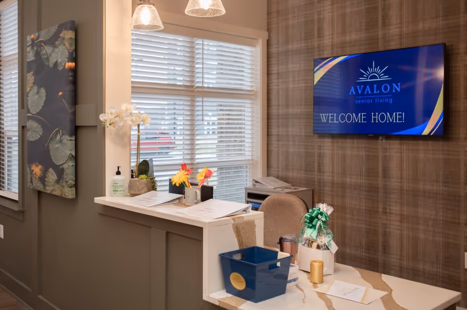 Reception area of Avalon Senior Living featuring a desk with documents, a blue basket, a gift basket with a green ribbon, a chair, and a wall-mounted TV screen displaying 'Avalon Senior Living WELCOME HOME!'. There are windows with blinds and decorative wall art on the side.
