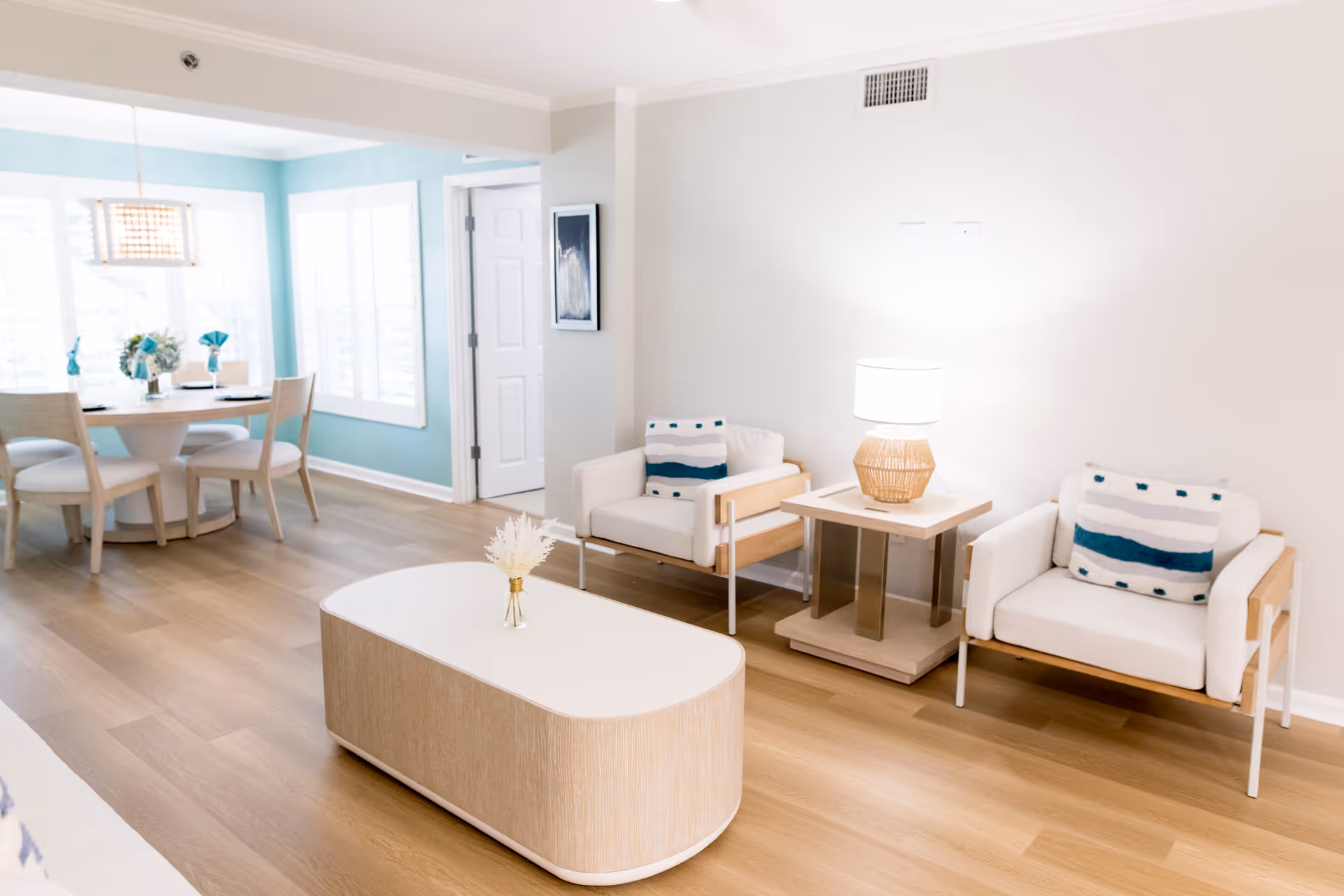 A bright and modern living room and dining area with light wood flooring. The living room features two white armchairs with wooden accents and patterned cushions, a small side table with a lamp, and a white oval coffee table with a small decorative vase. The dining area has a round table with four chairs and a hanging light fixture, set against a turquoise accent wall with large windows covered by white blinds.