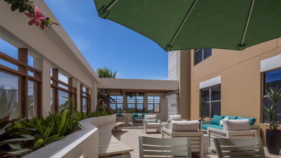 Outdoor patio area at Belmont Village Senior Living Fort Lauderdale with modern white seating furniture, green cushions, potted plants, a curved built-in bench with greenery, and a large green umbrella under a clear blue sky.