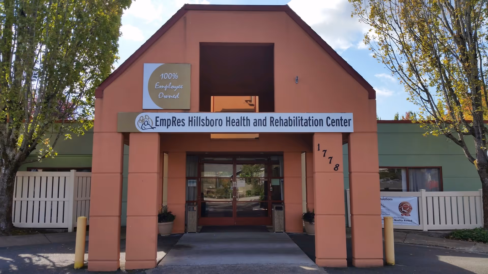 Front entrance of EmpRes Hillsboro Health and Rehabilitation Center with a peach-colored archway and a sign displaying the facility's name. There are trees on both sides and a clear sky in the background.
