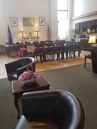 A spacious dining area with a long table covered with a red tablecloth and surrounded by wooden chairs. In the foreground, there are two black leather armchairs and a small wooden table with a basket of purple flowers. The background features a fireplace with lamps, framed pictures, an American flag, and a kitchen area with dark cabinets and appliances.