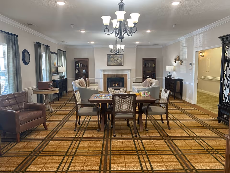 A cozy common area with a fireplace at the center back wall, flanked by two bookshelves. In front of the fireplace are two armchairs and a small table with a puzzle and board games. The room has patterned carpet flooring, two chandeliers hanging from the ceiling, and windows with curtains on the left side. There is a leather armchair on the left and a cabinet with a lamp on the right side of the room.
