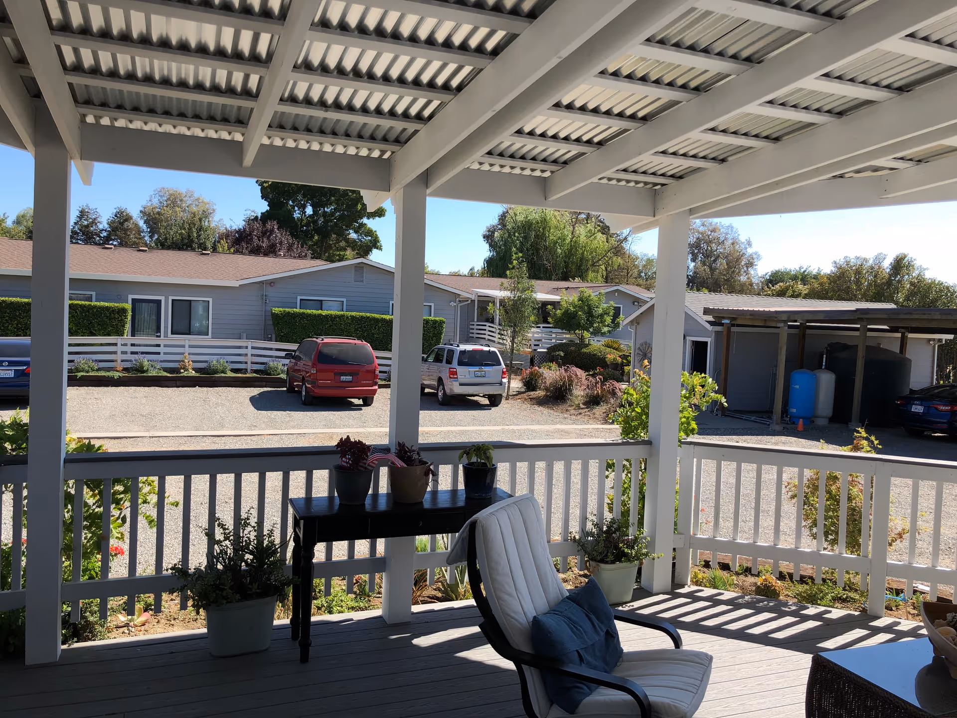 View from a covered porch with white railing and a cushioned chair with a blue pillow. There is a small black table with potted plants on it and additional plants on the porch floor. Beyond the porch, there is a gravel parking area with several parked cars and single-story residential buildings surrounded by greenery and trees under a clear blue sky.