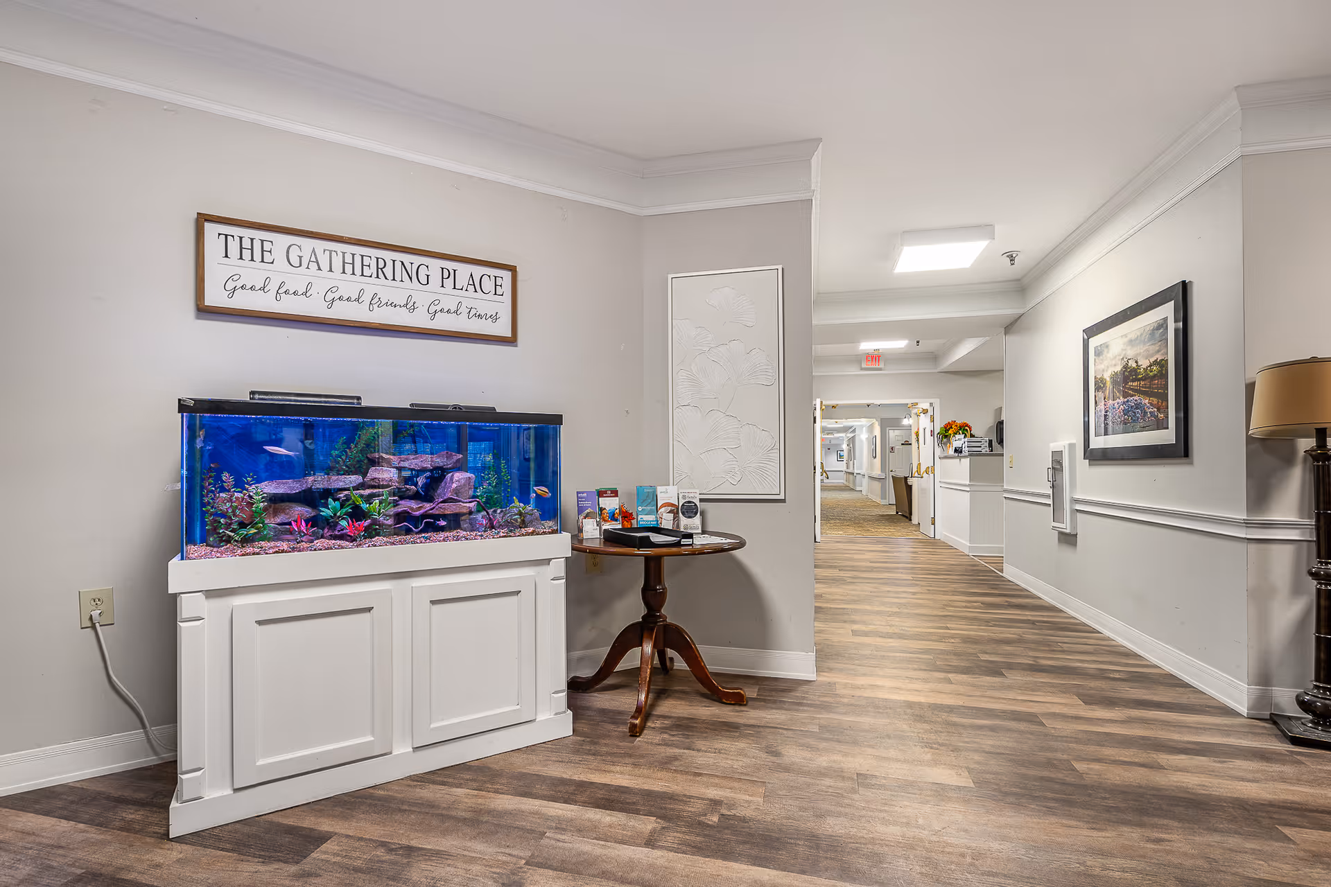 Interior hallway of a senior living facility with wood flooring, a large fish tank on a white stand, a round wooden table with brochures, a floor lamp, and framed artwork on the walls. A sign above the fish tank reads 'THE GATHERING PLACE Good food. Good friends. Good times.'