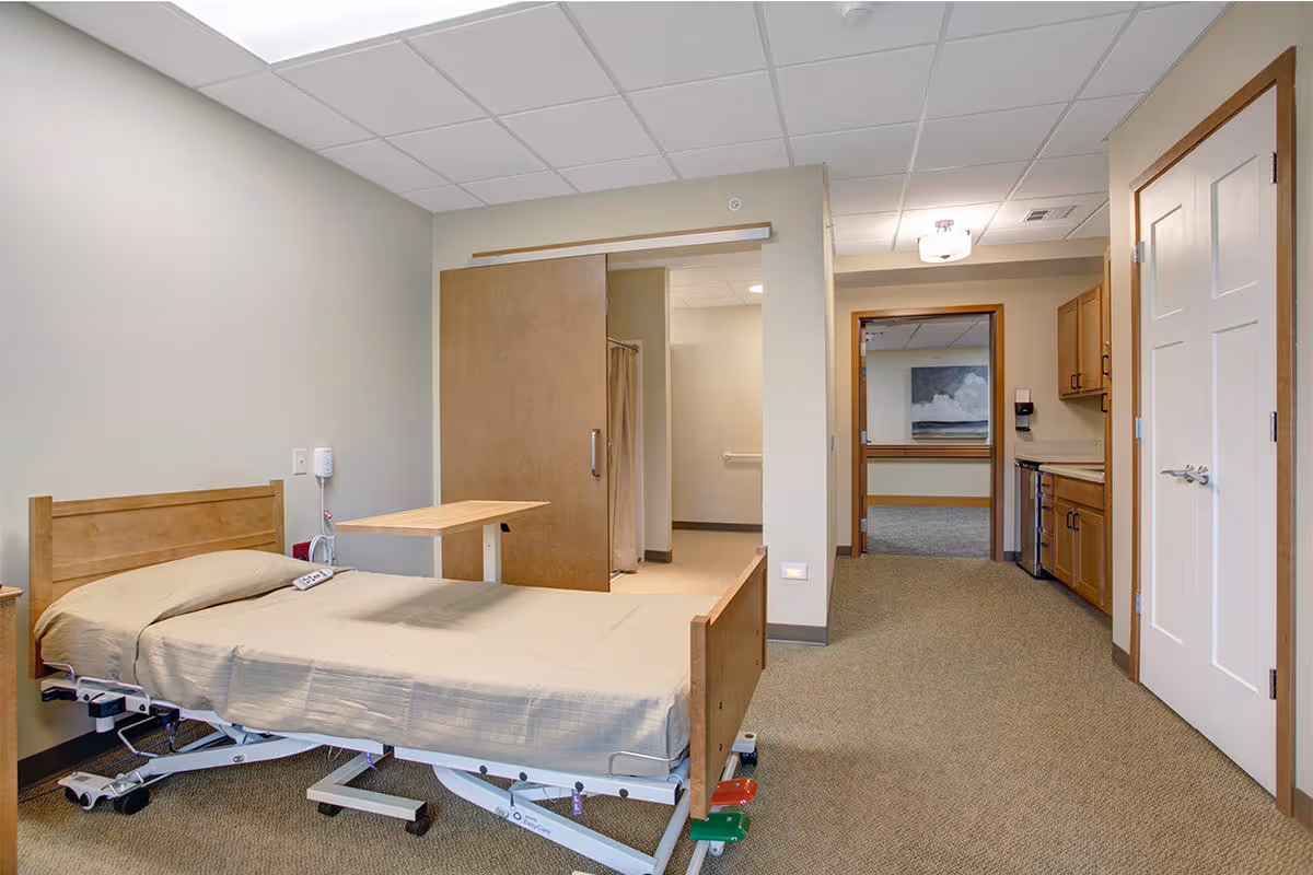 A senior living facility room at Prairie Gate featuring a hospital-style adjustable bed with beige bedding, a small overbed table, a sliding wooden door leading to a bathroom area with a shower curtain, and a kitchenette with wooden cabinets and a mini fridge. The room has beige walls, carpeted flooring, and a ceiling light fixture.