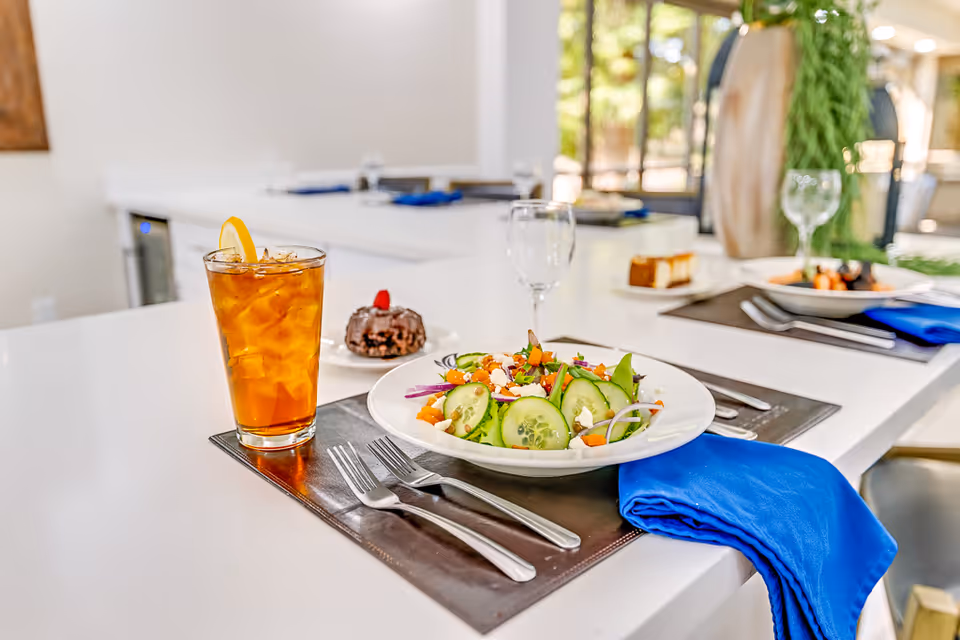 A plated salad with iced tea and dessert arranged on a white countertop with place settings and blue napkins in a dining area.