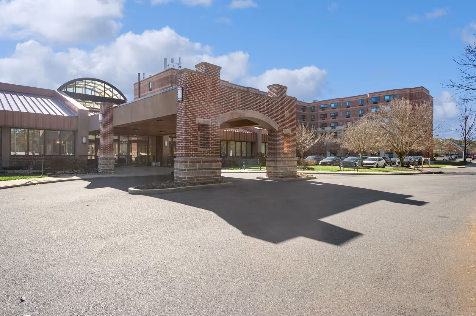 Front entrance of a brick senior living facility with a covered porte-cochère, parking lot, and multi-story building in the background.