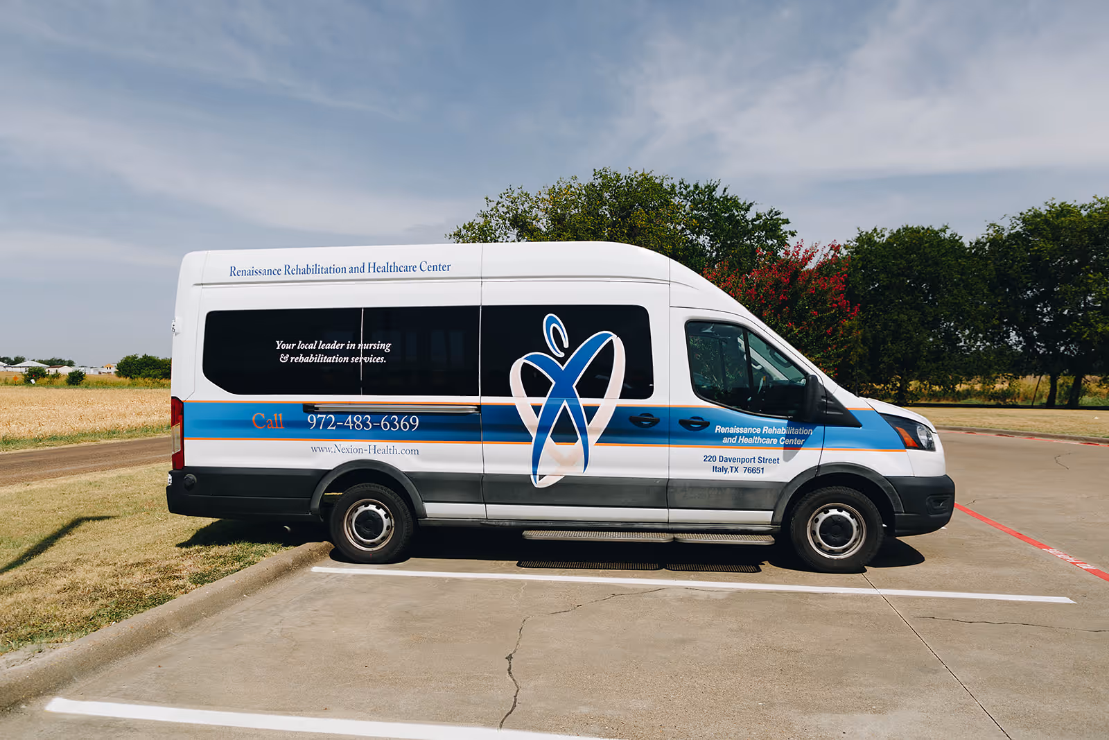 A white and blue shuttle van parked in a parking lot with the branding of Renaissance Rehabilitation and Healthcare Center. The van displays the center's name, contact number, website, and address on its side. Trees and a grassy area are visible in the background under a partly cloudy sky.
