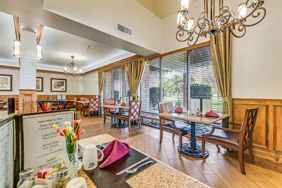 A bright and elegant dining room with wooden floors and large windows covered with blinds and curtains. Several tables are set with menus, folded napkins, and cutlery. The room features chandeliers hanging from the ceiling and framed artwork on the walls. Outside the windows, greenery and trees are visible.