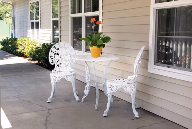White ornate metal bistro table and two matching chairs with a potted orange-flowered plant on the table set on a concrete porch beside a beige-sided building with windows.