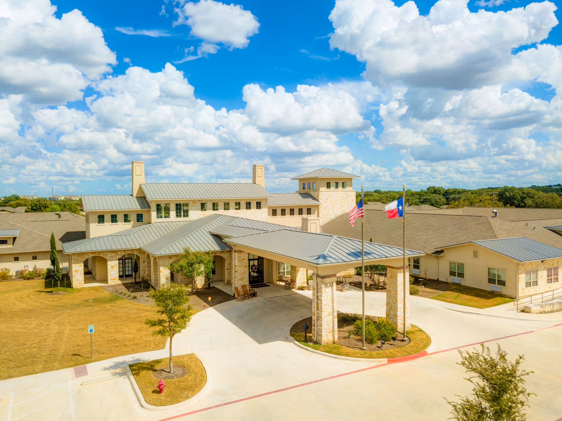 Front exterior of a large senior living facility with a covered porte-cochere, stone facade and flagpoles flying the American and Texas flags under a blue sky.