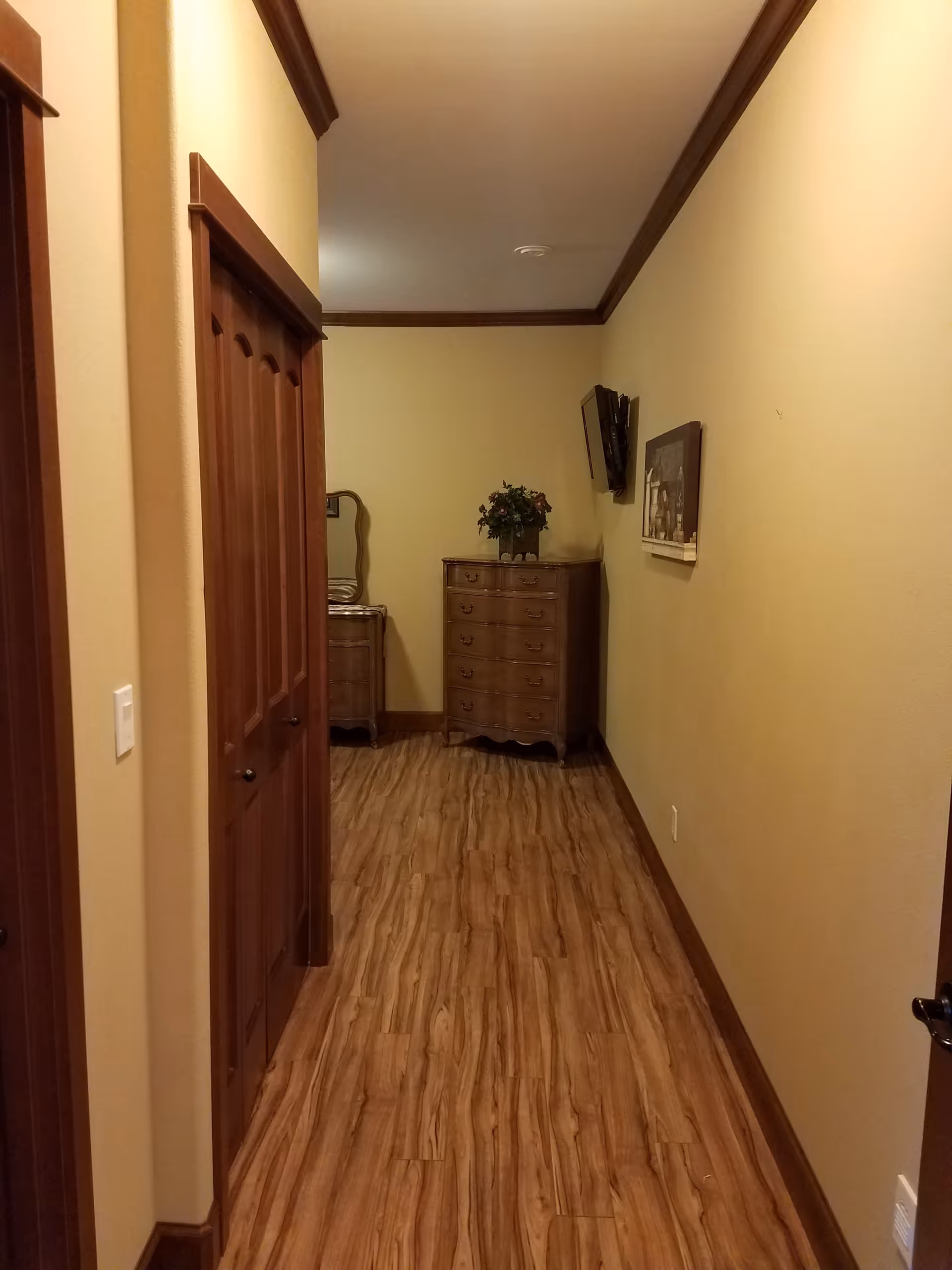 A narrow hallway with wooden flooring and beige walls. On the left side, there are wooden double doors. At the end of the hallway, there is a wooden dresser with a flower arrangement on top and a mirror partially visible behind it. A flat-screen TV is mounted on the right wall above a framed picture.