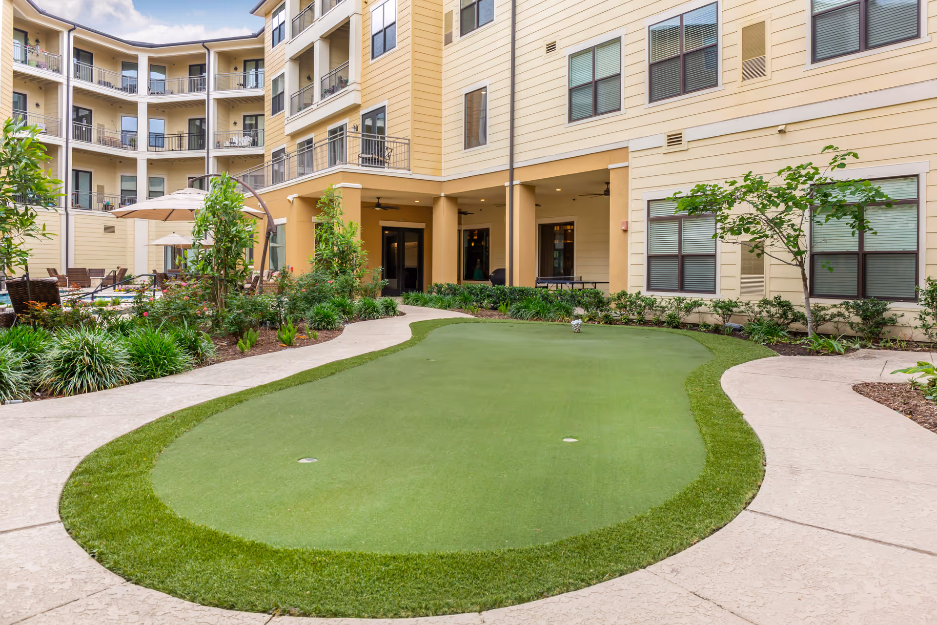 Outdoor courtyard area of a senior living facility featuring a putting green surrounded by a curved concrete walkway, landscaped plants, and a multi-story building with balconies and windows in the background.