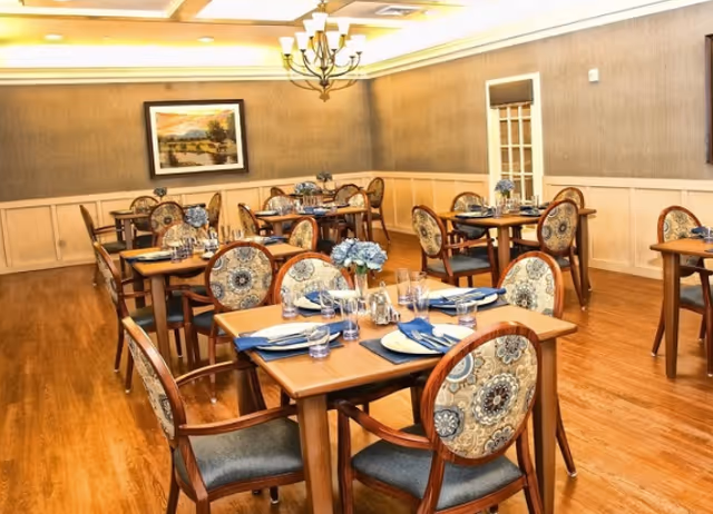 A well-lit dining room with multiple wooden tables and chairs arranged neatly. Each table is set with blue napkins, plates, glasses, and silverware. There are small flower arrangements on each table. The room has wooden flooring, beige walls with wainscoting, a chandelier hanging from the ceiling, and a framed landscape painting on the wall.