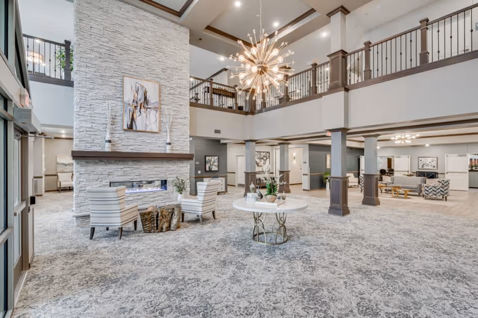 Spacious two-story senior living lobby with seating, a stone fireplace, central round table, and a large chandelier.