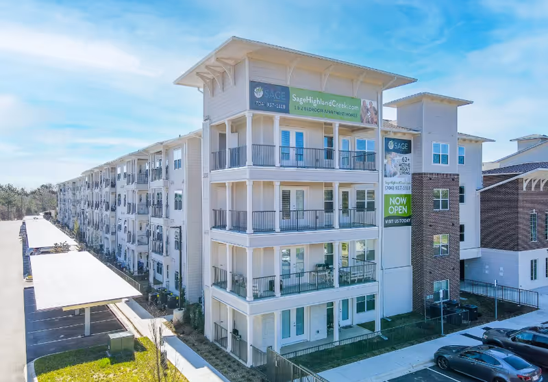 Exterior view of a multi-story Sage Highland Creek senior living apartment building with balconies and signage reading 'Now Open'.