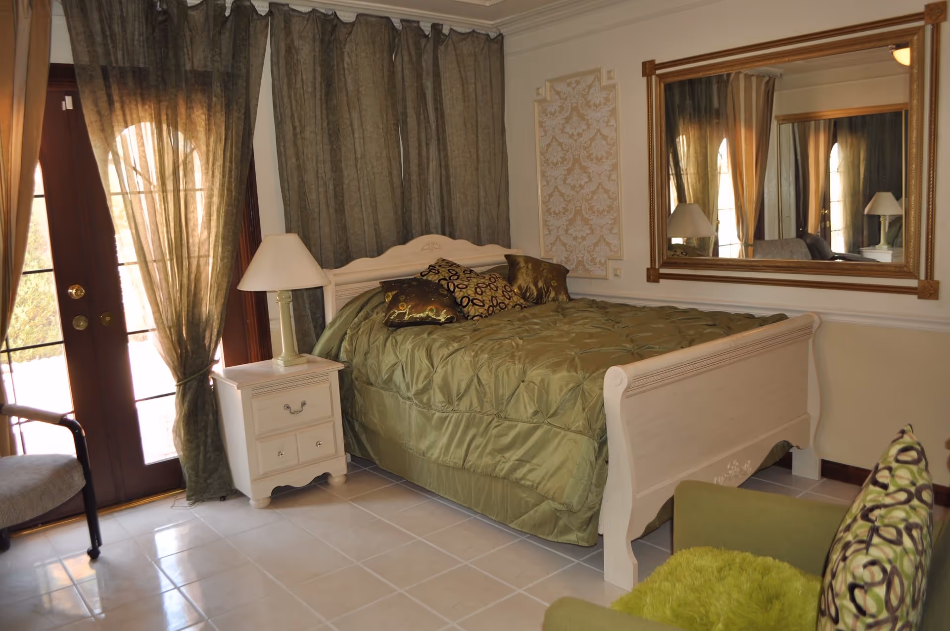 Bedroom with a green comforter on a white sleigh bed, nightstand and lamp, large mirror, and curtained French doors.