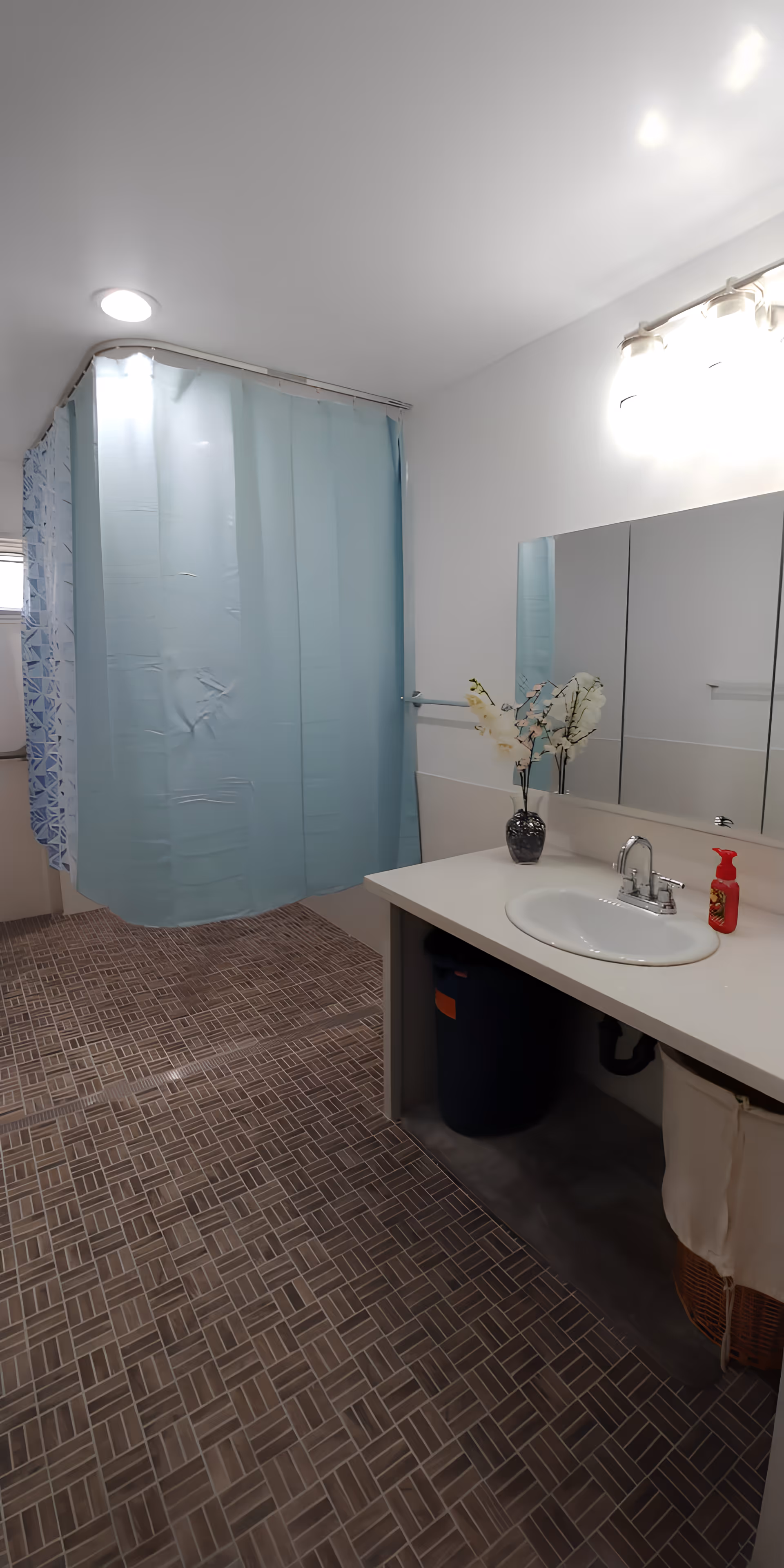 A clean bathroom with a blue shower curtain, a white sink with a silver faucet, a large mirror above the sink, a vase with white flowers on the countertop, and a red soap dispenser. The floor has brown patterned tiles and the walls are white.