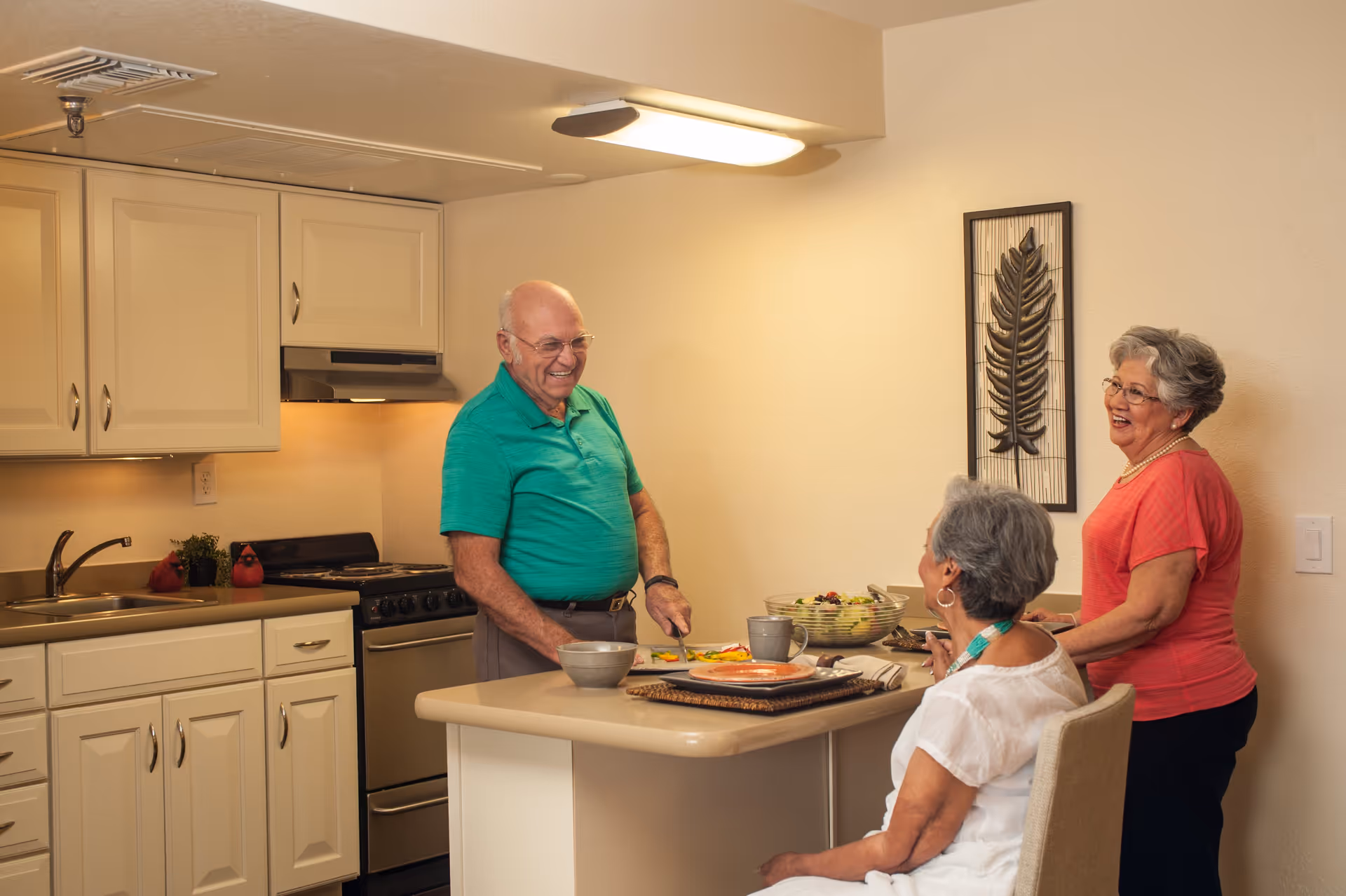 Three elderly people in a kitchen area. One man in a green shirt is standing behind a kitchen island, cutting vegetables. Two women, one seated and one standing, are engaged in conversation with him. The kitchen has cream-colored cabinets, a stove, and a sink. A decorative wall piece with a leaf design is visible on the wall.