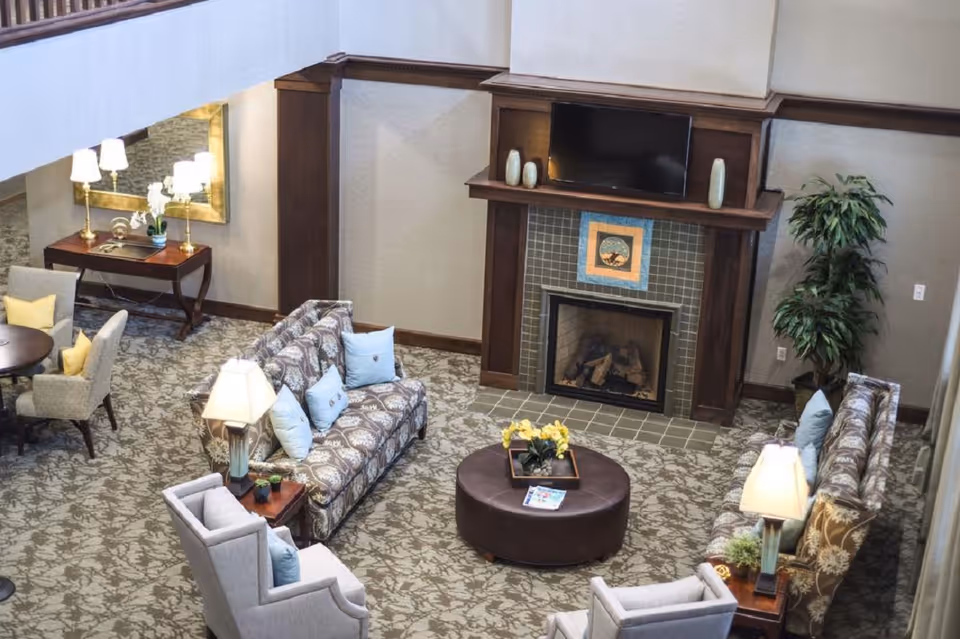 Spacious senior living lounge with patterned sofas and armchairs arranged around a round ottoman facing a fireplace and wall-mounted TV.