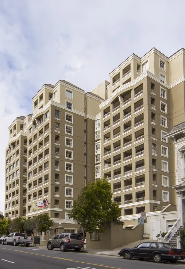 Exterior view of a multi-story residential building with balconies, parked cars along the street, and a tree in front. The building is beige with white trim and has an American flag and another flag near the entrance.
