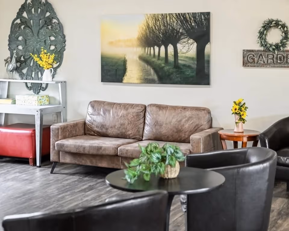 A cozy living room area with a brown leather sofa, two black leather armchairs, and a round black coffee table with a green potted plant. On the wall above the sofa is a large framed picture of a tree-lined path in a misty setting. To the left, there is a white shelf with decorative items including a vase with yellow flowers and a large ornate wall decoration. To the right, there is a small wooden side table with a vase of yellow flowers and a wall decoration with a wreath and the word 'GARDEN'. The floor is wood with a grayish tone.