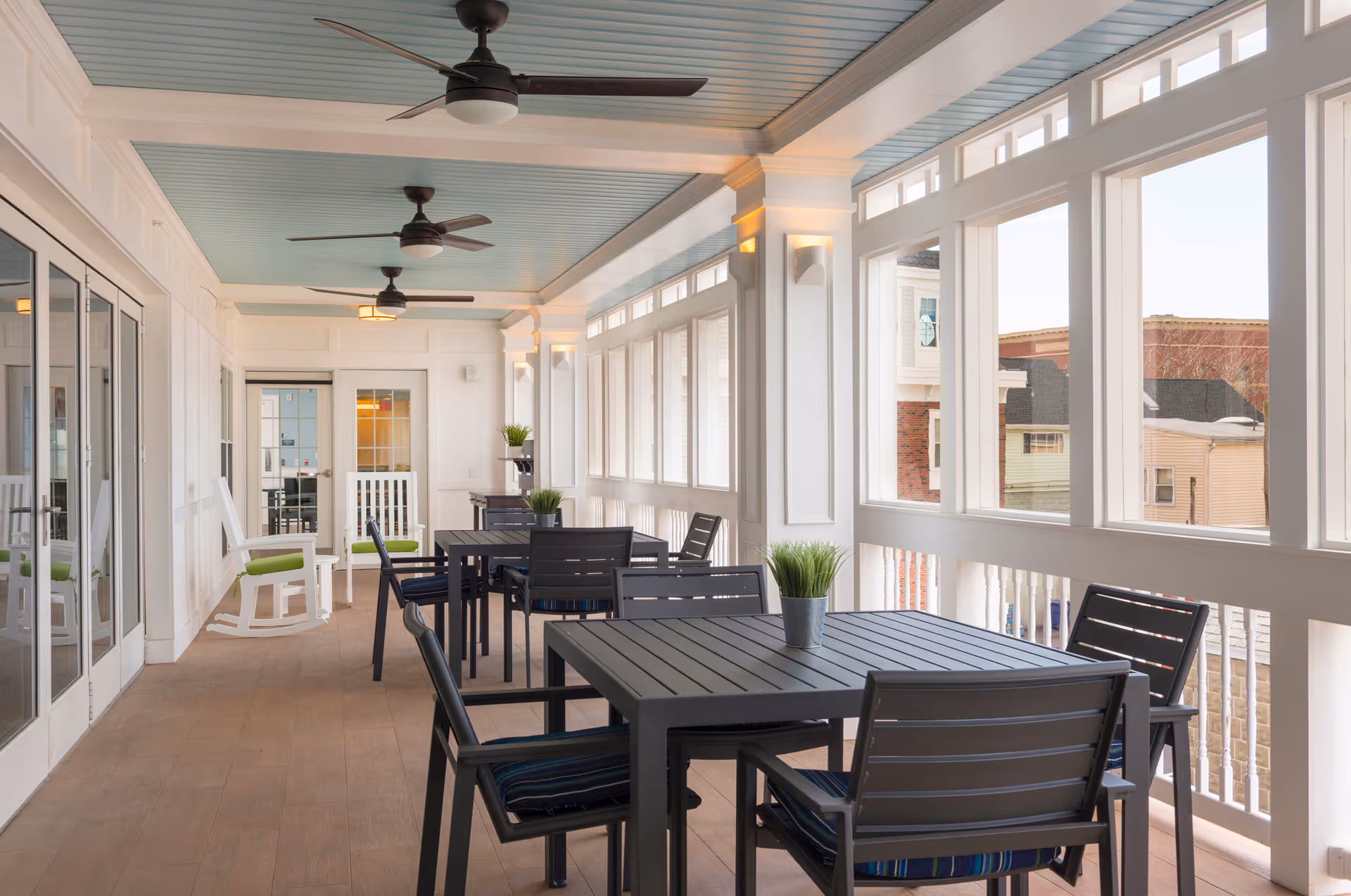 A bright and airy enclosed porch area with multiple black tables and chairs, each table decorated with a small potted plant. The space features white walls, large windows letting in natural light, ceiling fans, and a light blue ceiling. There are also white rocking chairs along one side.