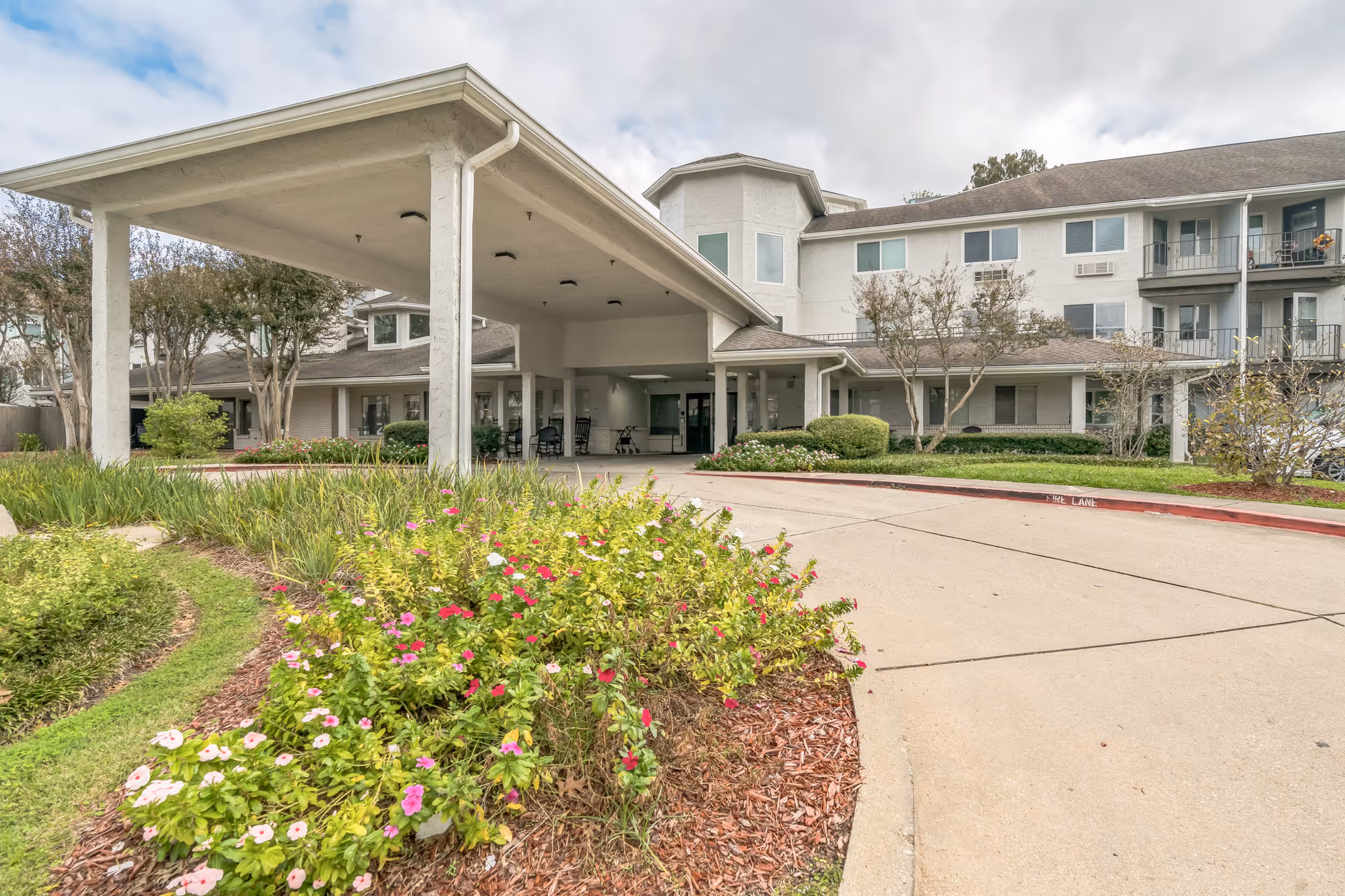 Front entrance of a multi-story senior living building with a covered porte-cochère, driveway, balconies, and landscaped flower beds.