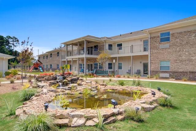 Outdoor view of a senior living facility named Prema at Suwanee Creek featuring a landscaped garden with a small pond surrounded by rocks and plants, a two-story building with balconies and windows, and a playground with a slide in the background under a clear blue sky.