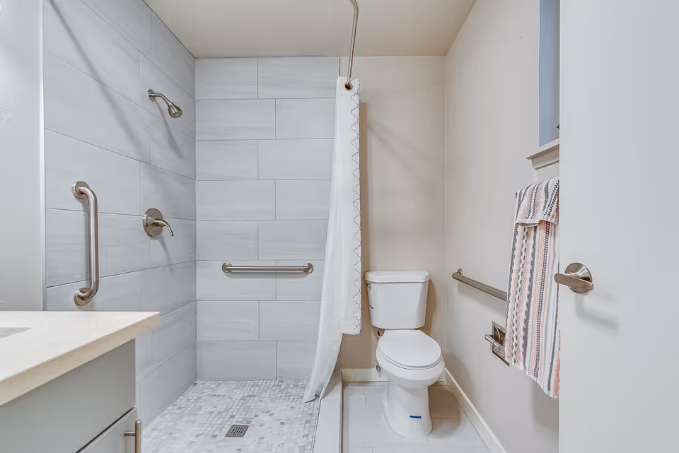 A clean, modern bathroom featuring a walk-in shower with a handheld showerhead and grab bars, a white toilet, a towel rack with a striped towel, and a light-colored countertop with a sink.