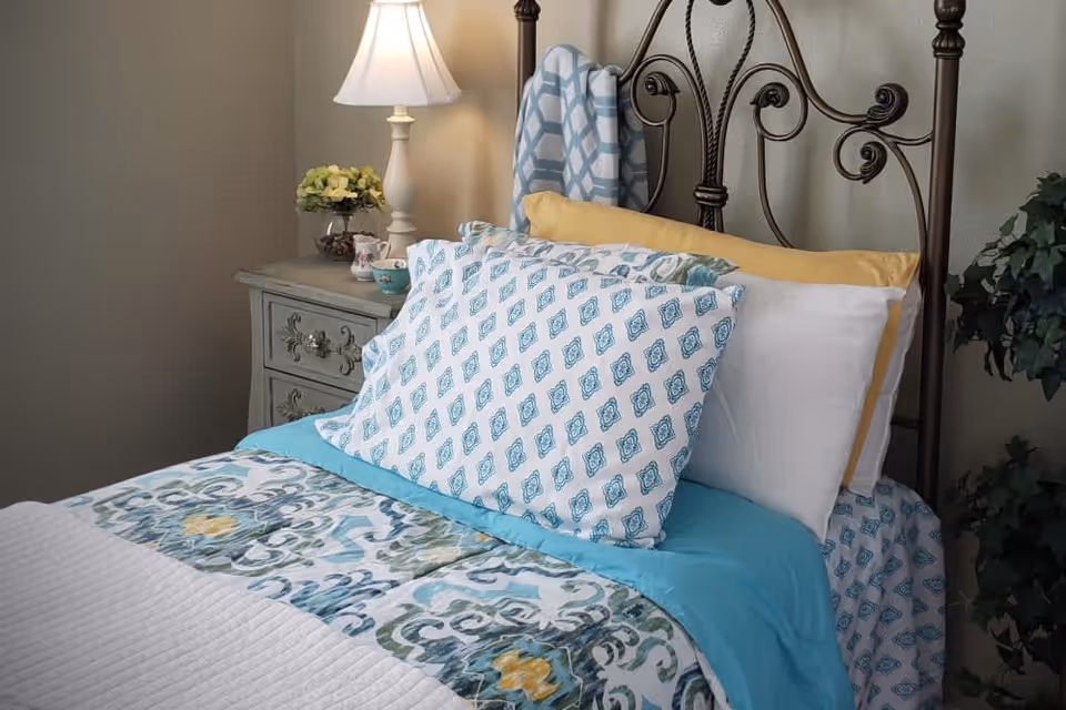 A neatly made bed with patterned blue and white pillows and a blue and white bedspread with yellow accents. The bed has a decorative metal headboard. Next to the bed is a vintage-style nightstand with a white table lamp, a small flower arrangement, and a teacup. A green plant is visible on the right side of the image.