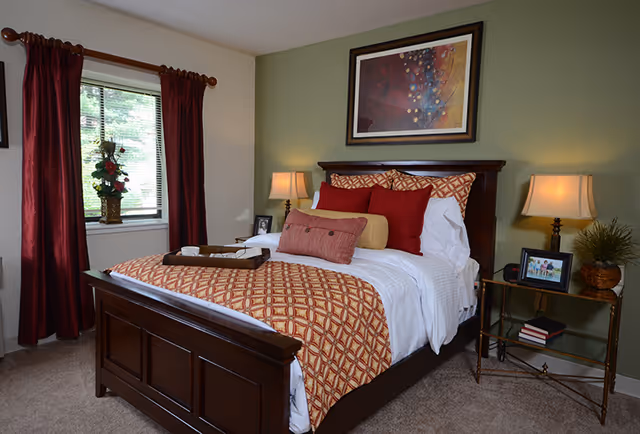 A cozy bedroom with a dark wooden bed frame, neatly made bed with white sheets and patterned orange and red pillows and blanket. Two bedside tables with lamps, a framed picture, books, and a plant. A window with red curtains and a vase with flowers is on the left side. A colorful abstract painting hangs on the green accent wall behind the bed.