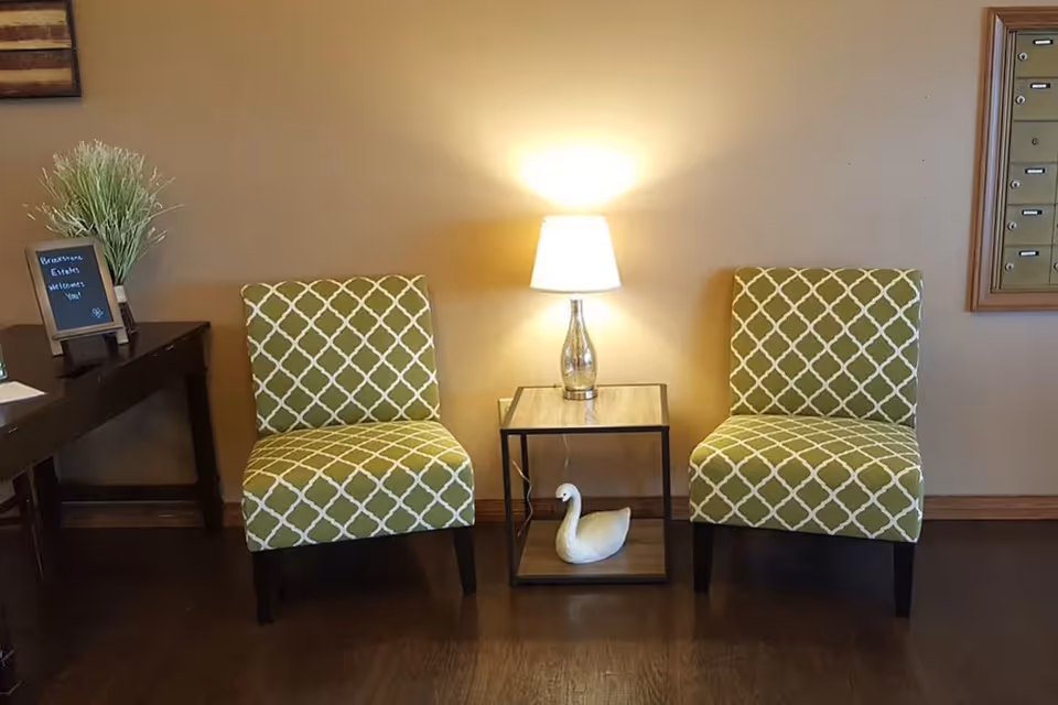 Two green patterned chairs flank a small side table with a lamp and a decorative swan in a facility lobby.