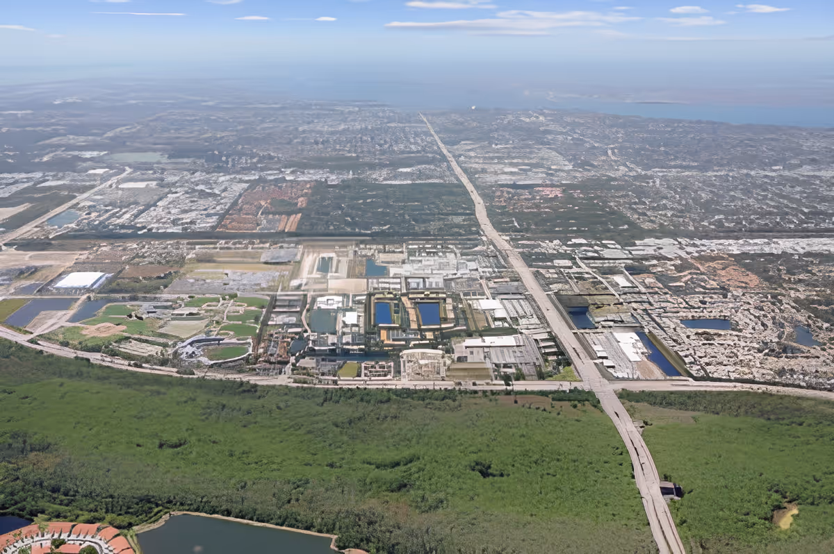 Aerial view of a large urban area with a mix of industrial, residential, and green spaces. A major road runs vertically through the center of the image, with various buildings and water bodies visible. The foreground shows dense green vegetation and a small body of water.