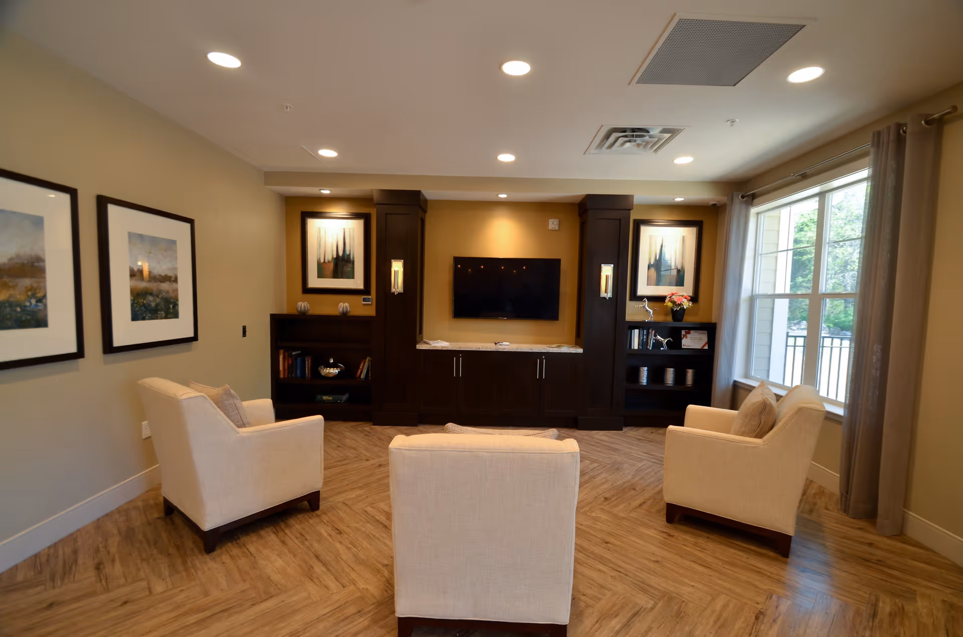 A cozy living room area with three beige armchairs arranged around a wall-mounted flat-screen TV. The room features wooden flooring, built-in dark wood cabinetry with shelves on either side of the TV, framed artwork on the walls, and a large window with curtains letting in natural light.