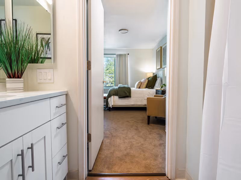 View from a bathroom looking into a bedroom in a senior living facility. The bathroom has a white vanity with drawers and a green plant on the countertop. The bedroom features a bed with white bedding and green pillows, a window with curtains, a beige armchair, and framed artwork on the wall.