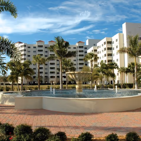 Front view of a multi-story white senior living building with palm trees and a large fountain in the foreground under a blue sky.