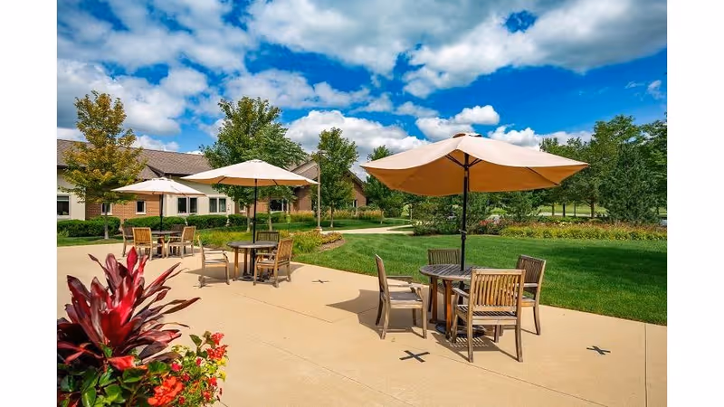 Outdoor patio area with round tables and wooden chairs under large beige umbrellas, surrounded by green grass, trees, and a partly cloudy blue sky. A building is visible in the background.