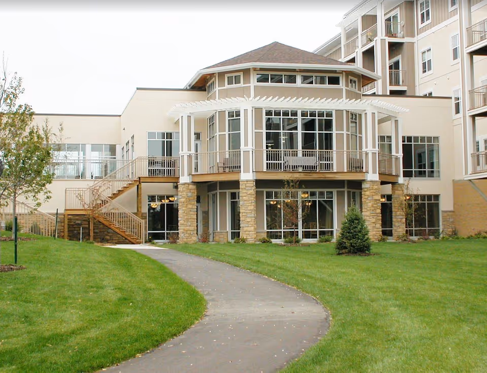 Front exterior of a multi-story senior living building with large windows, a covered second-story balcony and a paved walkway across a green lawn.