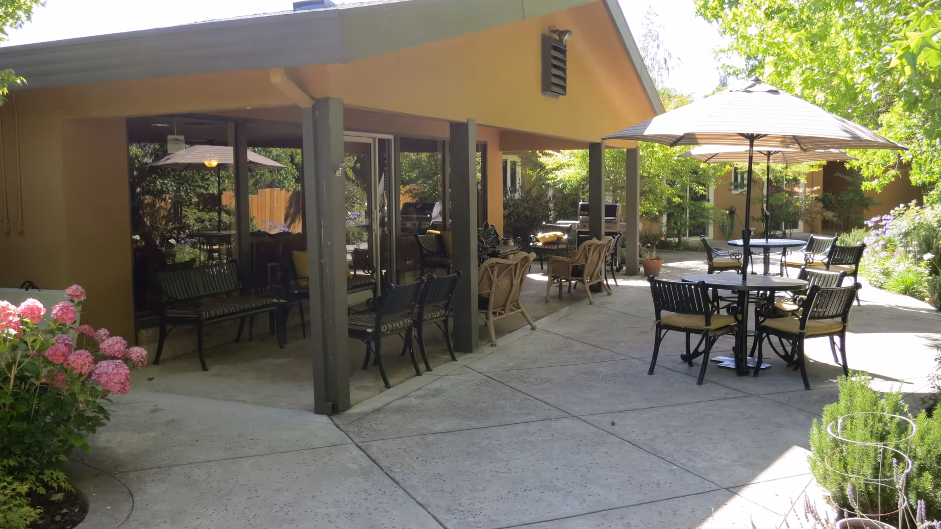 Outdoor patio area with round tables, umbrellas, and various seating under a covered walkway beside a building, surrounded by plants.