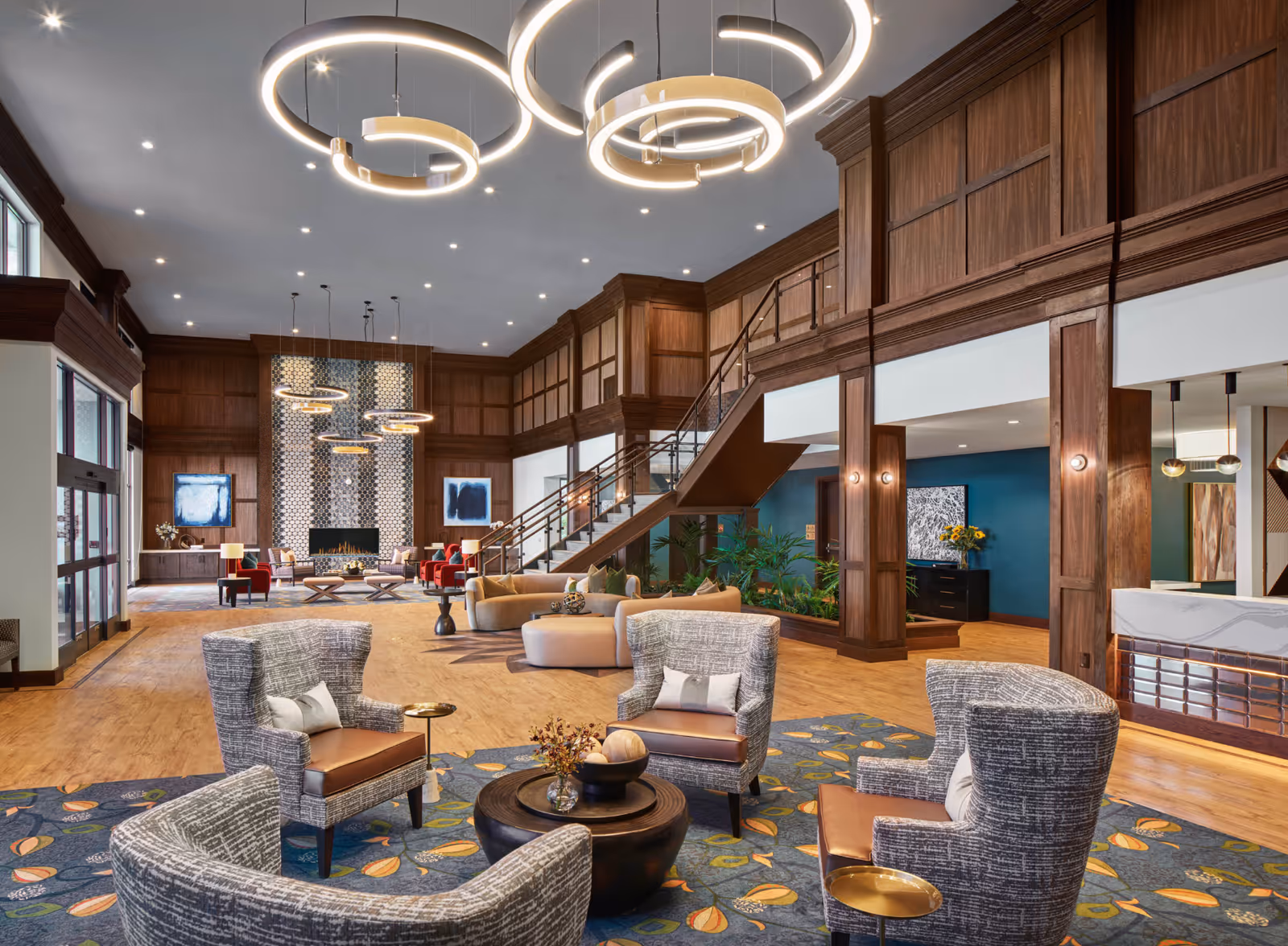 Spacious and elegant senior living facility lobby with modern circular pendant lights, wood-paneled walls, a fireplace with mosaic tile surround, comfortable seating areas including armchairs and sofas, a staircase leading to an upper level, and decorative plants.
