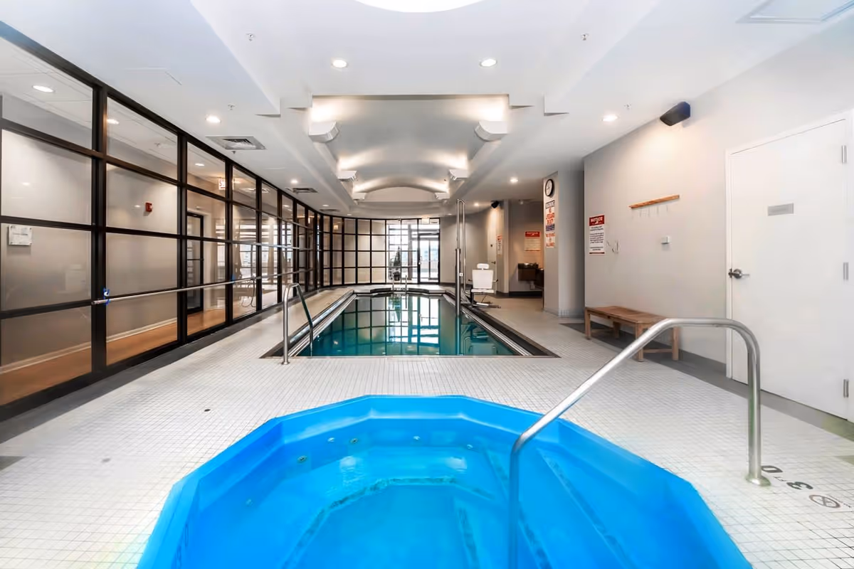 Indoor pool area with a rectangular swimming pool and a blue hot tub in the foreground. The room has white tiled floors and walls, a ceiling with recessed lighting, and large glass windows on one side. There is a bench and some hooks on the wall to the right, along with safety and notice signs.