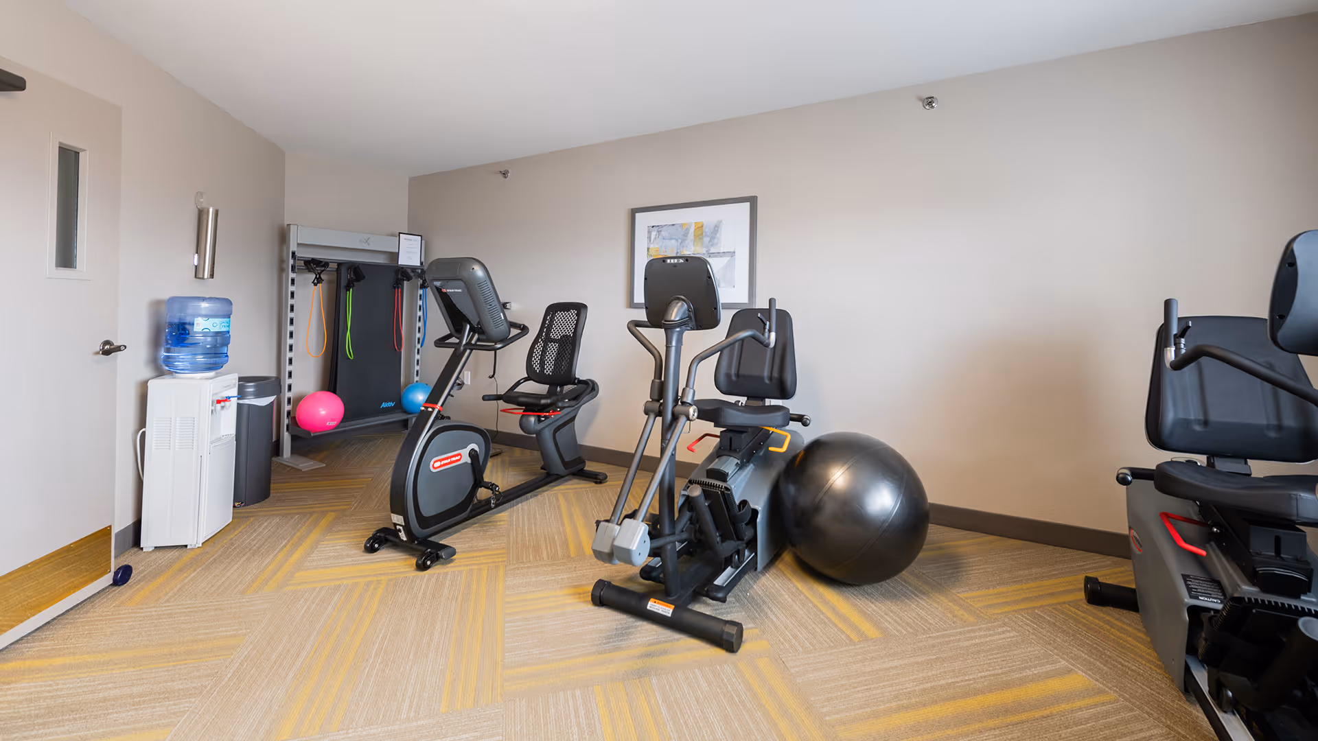 A small fitness room with exercise equipment including two recumbent stationary bikes, an elliptical machine, a large exercise ball, resistance bands hanging on a rack, and a water cooler near the door. The room has beige walls and patterned carpet flooring.