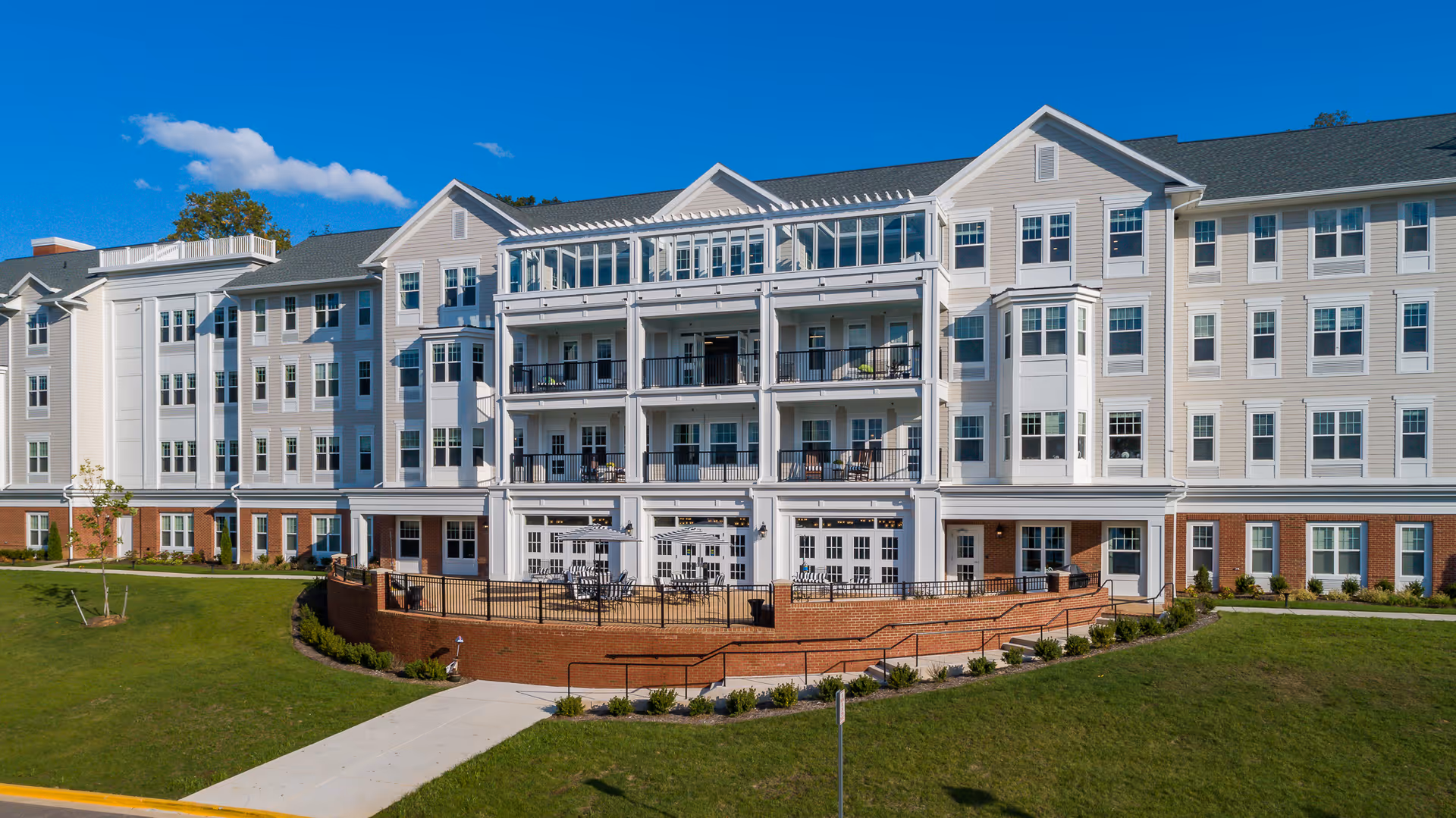 Exterior view of a large, multi-story senior living facility building with white siding and brick accents. The building features multiple balconies and large windows, with a patio area in front that has tables and chairs. The sky is clear and blue with a few clouds.
