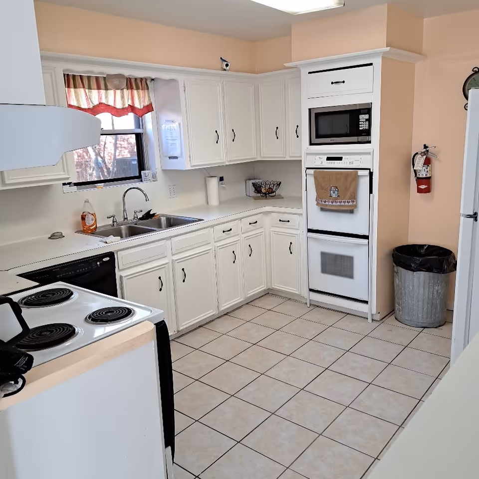 Bright white kitchen with tiled floor, white cabinets and countertops, a sink under a window, stove, microwave/oven stack, and a trash can.