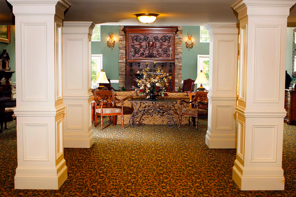 Interior view of a senior living facility lounge area with ornate white columns, patterned carpet, a decorative wooden fireplace, two table lamps, wooden chairs, and a floral arrangement on a wrought iron table.