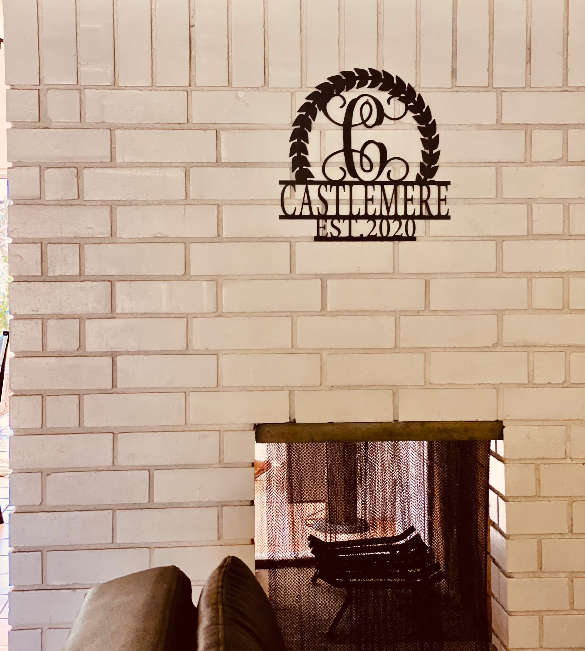 A white brick wall with a metal decorative sign that reads 'Castlemere EST 2020' mounted above a fireplace with a mesh screen. Part of a brown cushioned chair is visible in front of the fireplace.