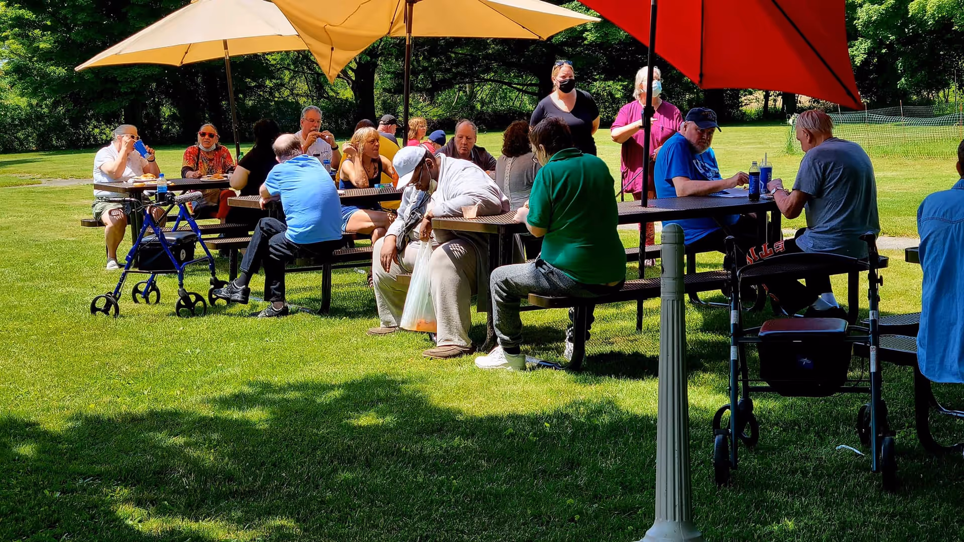 A group of elderly people sitting and socializing at picnic tables under large umbrellas in a grassy outdoor area with trees in the background. Some individuals are wearing masks and there are walkers nearby.